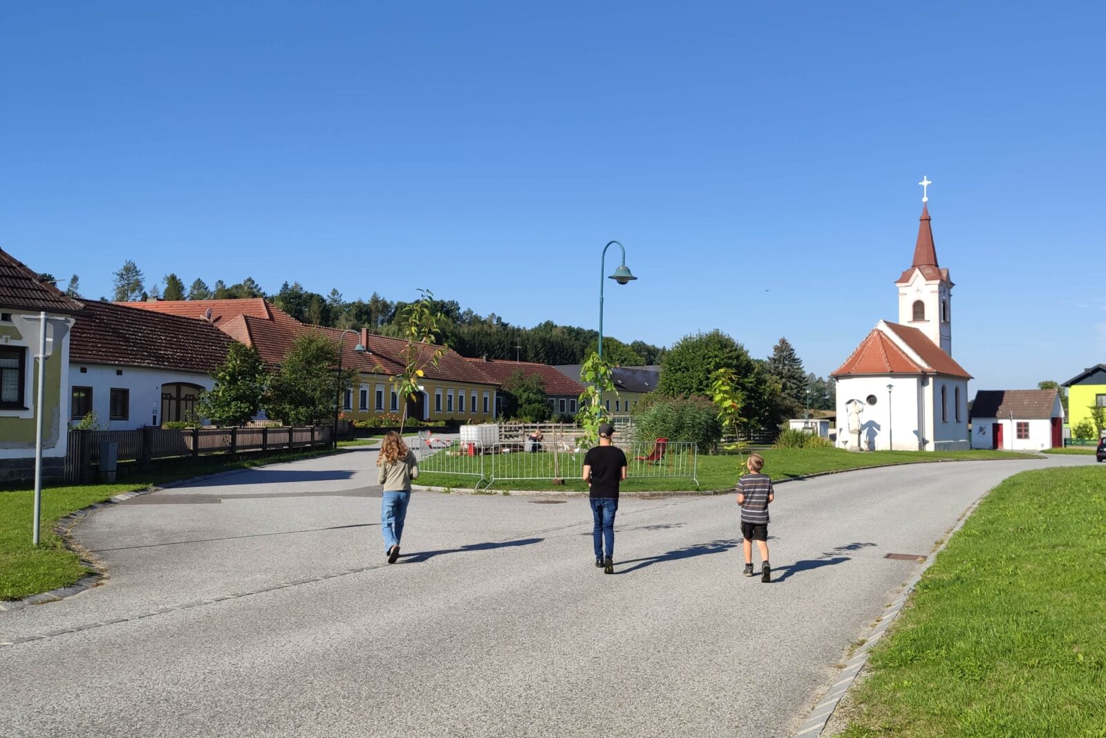Drei Menschen tragen Pflanzen über den Ortskern von Rafings. Im Hintergrund befindet sich die Dorfkirche und der blaue Himmel.
